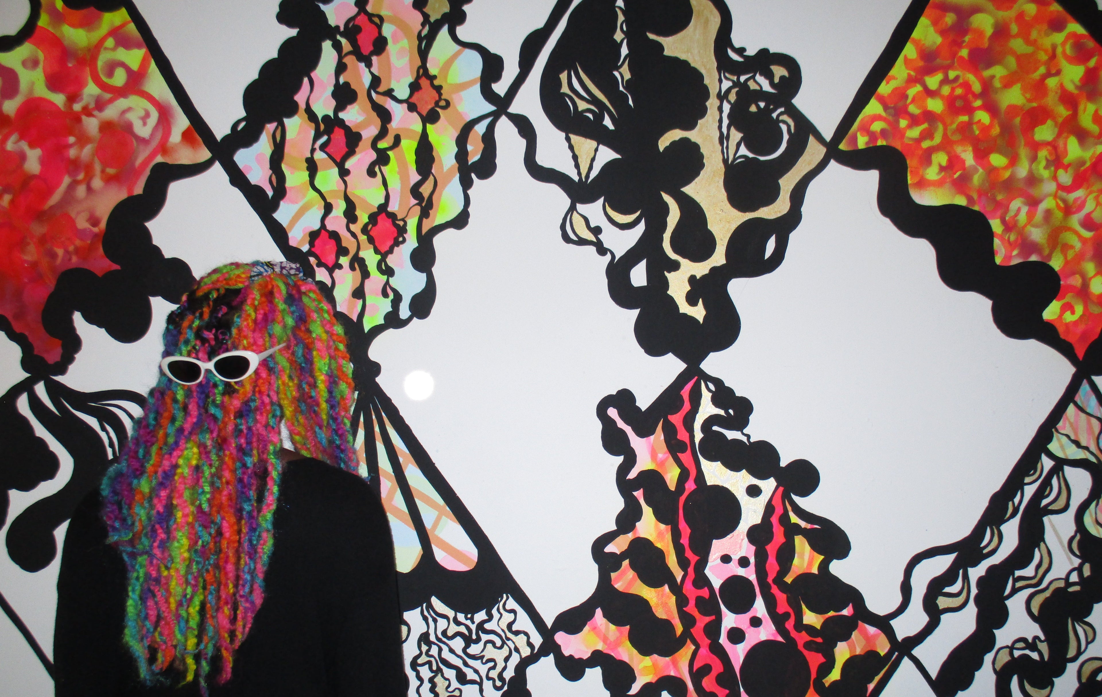 Jasmin Issaka with colourful psychedelic hair and sunglasses standing in front of her stylised abstract psychedelic mural design.