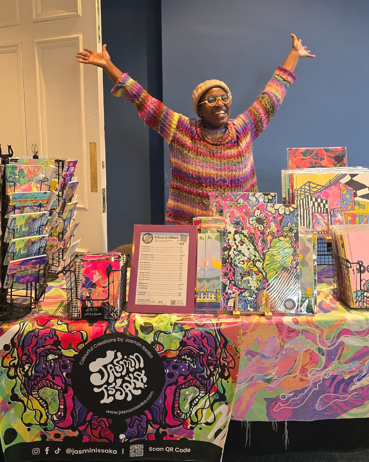 Afro-Psychedelic Artist, Jasmin Issaka, at her Market Stall. Colourful art display with various pieces of art prints, psychedelic posters, stationary, cards on a market stall stand against a dark blue wall.
